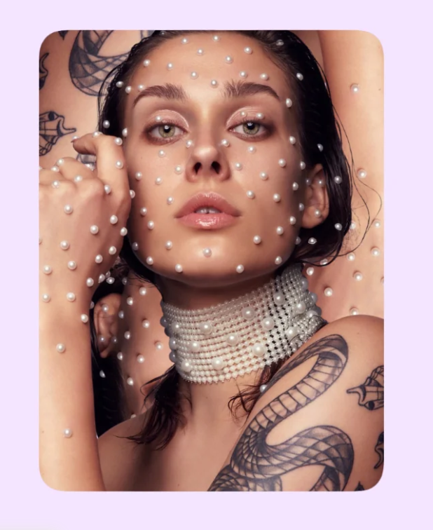 A close-up portrait of a woman with many small white pearls applied to her face, neck, and shoulders, including a pearl choker. She has visible tattoos and is posed against a pale lavender background with a rounded square crop around her.